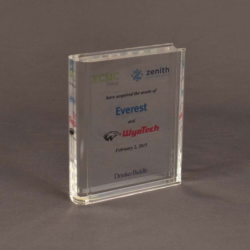Angled view of large slim book-shaped Lucite® embedment featuring corporate branding, acquisition details, and a sleek rectangular form